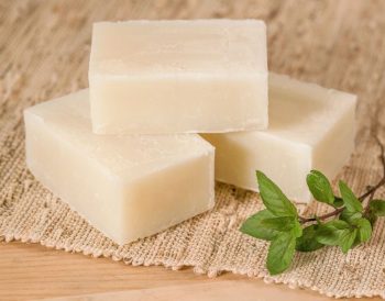 Mutfak tezgâhında eldivenli bir kişinin cam kapta yağları karıştırdığı, yanında kostik, su, silikon kalıplar ve bitkisel malzemelerin yer aldığı evde sabun yapımı hazırlık aşaması.