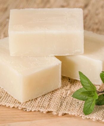 Mutfak tezgâhında eldivenli bir kişinin cam kapta yağları karıştırdığı, yanında kostik, su, silikon kalıplar ve bitkisel malzemelerin yer aldığı evde sabun yapımı hazırlık aşaması.