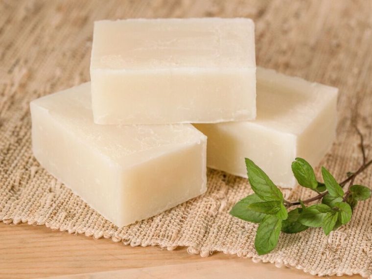 Mutfak tezgâhında eldivenli bir kişinin cam kapta yağları karıştırdığı, yanında kostik, su, silikon kalıplar ve bitkisel malzemelerin yer aldığı evde sabun yapımı hazırlık aşaması.