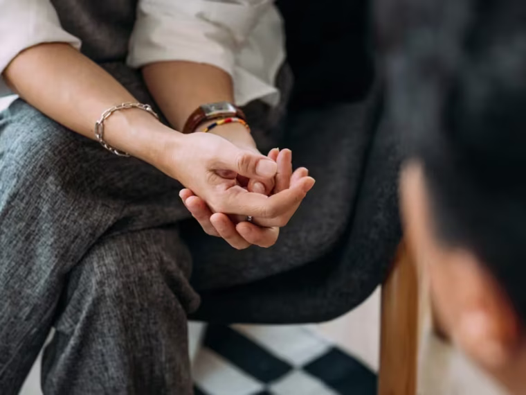 Bir çift el ele tutuşmuş, birbirlerine bakıyor; arka planda sakin bir ortam, güven ve yakınlık hissi yaratıyor.