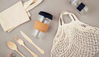 Mutfak tezgâhında yan yana duran cam saklama kapları, metal matara, bambu pipet, bez alışveriş çantası ve file içinde sebzelerin görüldüğü; arkada tezgâh üstünde tek kullanımlık plastik ürünlerin olmadığı sade bir ortam.