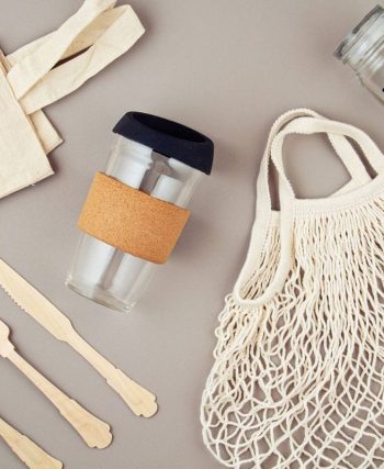 Mutfak tezgâhında yan yana duran cam saklama kapları, metal matara, bambu pipet, bez alışveriş çantası ve file içinde sebzelerin görüldüğü; arkada tezgâh üstünde tek kullanımlık plastik ürünlerin olmadığı sade bir ortam.