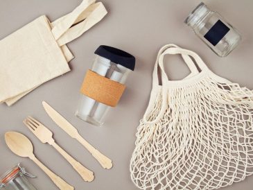 Mutfak tezgâhında yan yana duran cam saklama kapları, metal matara, bambu pipet, bez alışveriş çantası ve file içinde sebzelerin görüldüğü; arkada tezgâh üstünde tek kullanımlık plastik ürünlerin olmadığı sade bir ortam.