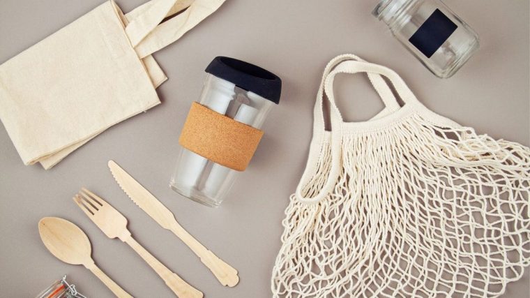 Mutfak tezgâhında yan yana duran cam saklama kapları, metal matara, bambu pipet, bez alışveriş çantası ve file içinde sebzelerin görüldüğü; arkada tezgâh üstünde tek kullanımlık plastik ürünlerin olmadığı sade bir ortam.